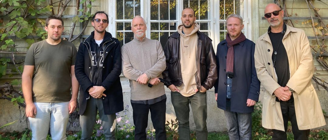 Fünf Männer stehen vor einem Haus mit einer Rankgitter, das mit Weinranken bedeckt ist. Sie tragen verschiedene Jacken und Hosen, und einer hat einen Schal. Das Haus hat große Fenster und eine Ziegelmauer.