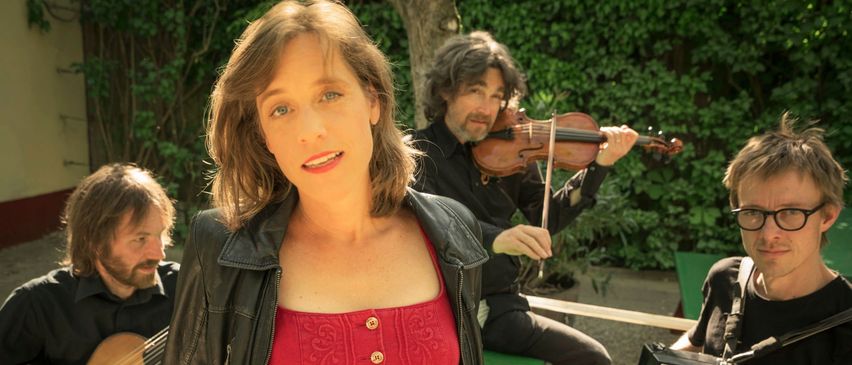 Eine Frau mit kurzen braunen Haaren steht vor einem sitzenden Geiger und Akkordeonspieler, alle in Schwarz gekleidet. Die Frau trägt ein rotes Shirt und eine schwarze Jacke. Hinter ihnen sind Grünpflanzen und eine Bank zu sehen.