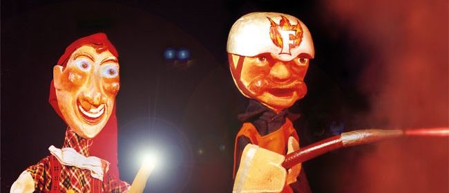 Zwei Comicfiguren in einer dunklen Umgebung, eine mit einer Taschenlampe und die andere mit einem Feuerwehrhelm und einer Axt, stehen vor einem roten Hintergrund.