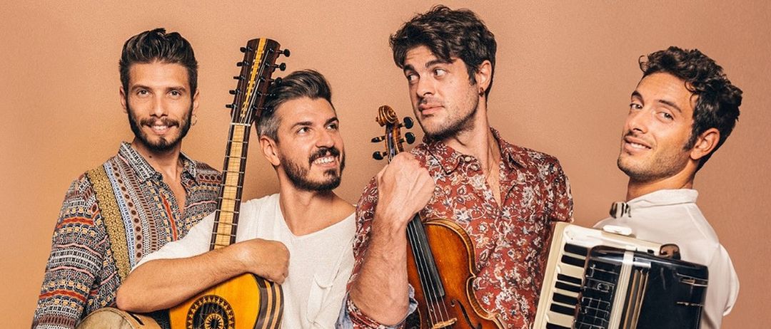 Fünf Männer posieren für ein Foto mit Musikinstrumenten. Ein Mann hält eine Gitarre, ein anderer eine Geige, ein dritter eine Rassel, während der Mann in der Mitte ein gemustertes Hemd trägt und der Mann rechts ein weißes Hemd trägt.