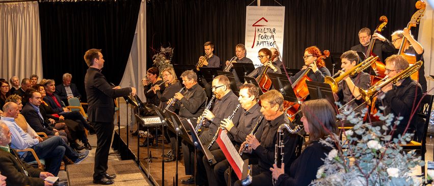 Eine Gruppe von Musikern tritt auf der Bühne auf, mit einem Banner dahinter, das 'Kultur Forum Amthor' steht. Der Dirigent führt mit einem Taktstock.