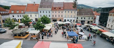 An aerial view of a town square filled with people and vendors. Colorful tents and awnings are set up, and cars are parked on the side. Trees line the street.