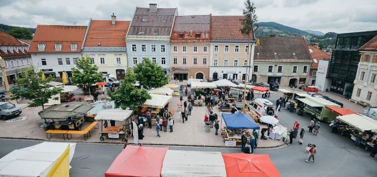 Ein Luftbild eines Stadtplatzes, gefüllt mit Menschen und Verkäufern. Bunte Zelte und Markisen sind aufgebaut, und Autos sind am Straßenrand geparkt. Bäume säumen die Straße.