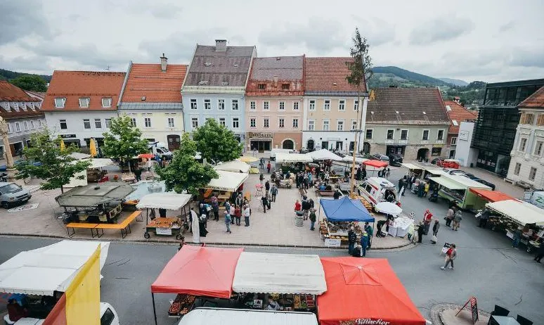 Ein Luftbild eines Stadtplatzes, gefüllt mit Menschen und Verkäufern. Bunte Zelte und Markisen sind aufgebaut, und Autos sind am Straßenrand geparkt. Bäume säumen die Straße.