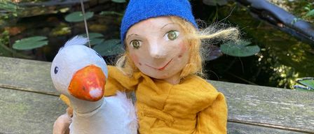 Eine Puppe in einem blauen Hut hält eine weiße Spielzeuggans mit orangefarbenem Schnabel. Die Puppe trägt ein gelbes Hemd und sitzt auf einer Bank am Teich mit Seerosenblättern.