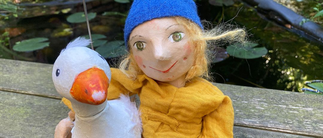 Eine Puppe in einem blauen Hut hält eine weiße Spielzeuggans mit orangefarbenem Schnabel. Die Puppe trägt ein gelbes Hemd und sitzt auf einer Bank am Teich mit Seerosenblättern.