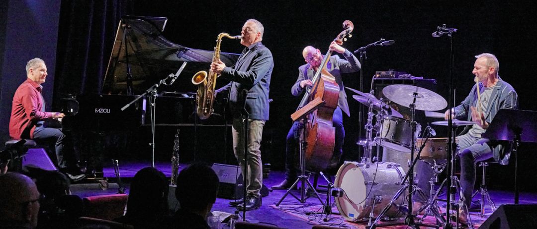 Eine Jazzband spielt auf der Bühne, mit einem Saxophonisten, Bassisten und Schlagzeuger. Ein Klavier und Mikrofone stehen dahinter. Das Publikum sitzt davor, beleuchtet von lila Lichtern.