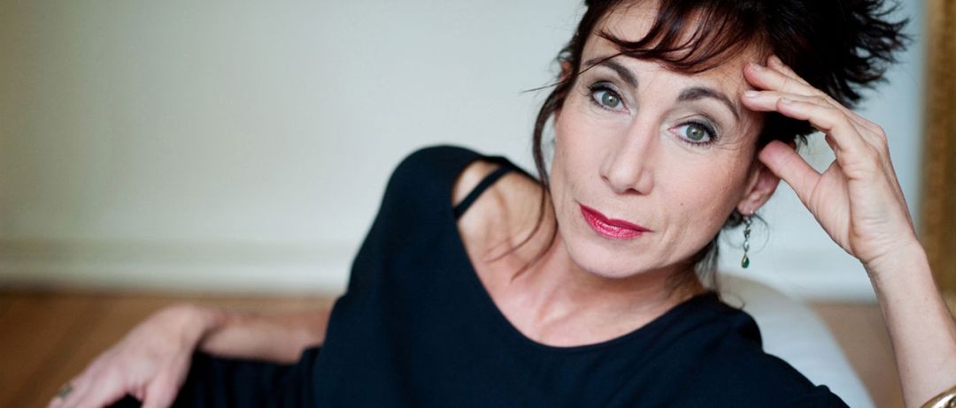 Eine Frau mit kurzen Haaren in einem schwarzen Kleid und rotem Lippenstift liegt auf dem Boden und stützt ihren Kopf in ihrer Hand. Sie hat einen ernsten Ausdruck und schaut nach vorne.