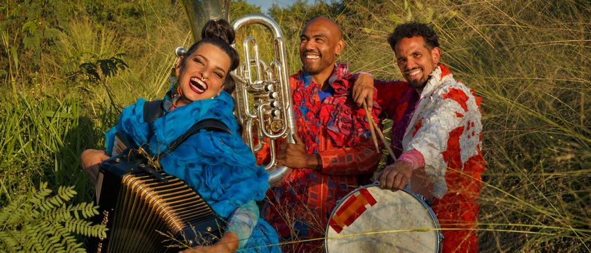 Drei Musiker in bunten Outfits stehen in einem Feld und lächeln. Eine Frau spielt Akkordeon, ein Mann hält eine Trommel und ein anderer Mann spielt Tuba.