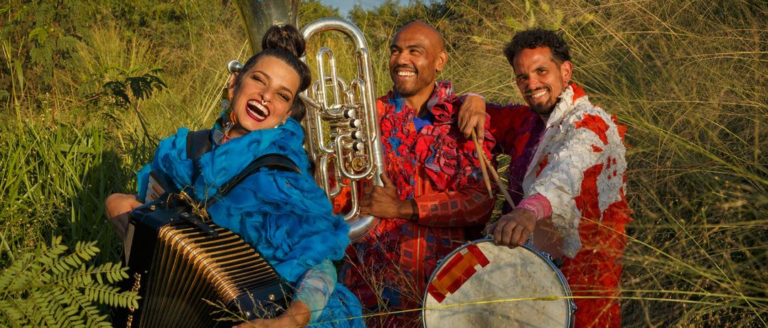 Drei Musiker in bunten Outfits stehen in einem Feld und lächeln. Eine Frau spielt Akkordeon, ein Mann hält eine Trommel und ein anderer Mann spielt Tuba.