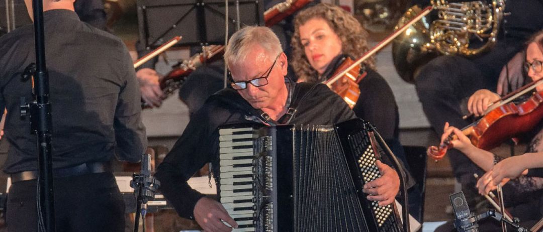 Ein Mann spielt Akkordeon in einem Konzert mit einem Streichquartett, einschließlich Geige, und Blechblasinstrumenten wie einer französischen Trompete.