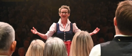 Eine Frau in traditioneller Kleidung und mit Brille steht auf der Bühne, lächelt und gestikuliert, während eine Menge Menschen sie beobachtet.