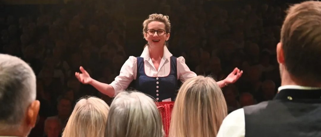 Eine Frau in traditioneller Kleidung und mit Brille steht auf der Bühne, lächelt und gestikuliert, während eine Menge Menschen sie beobachtet.
