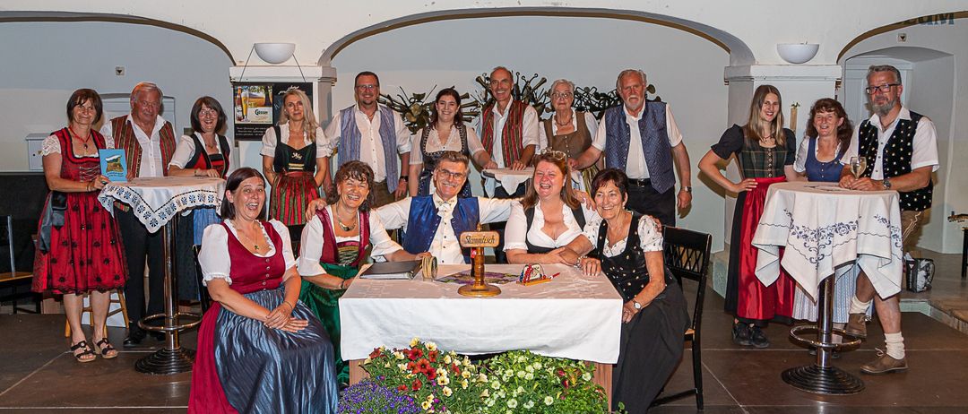 Eine Gruppe von Menschen in traditioneller deutscher Kleidung sitzt um einen Tisch mit Blumen herum und posiert für ein Foto. Der Tisch hat ein Holzschild mit der Aufschrift 'Stolz'. Der Raum ist gut beleuchtet.