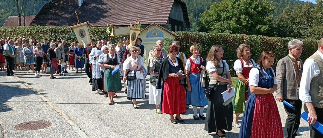 Eine Gruppe von Menschen in traditioneller Kleidung geht in einer Reihe vor einem Gebäude. Im Hintergrund befindet sich ein Haus mit einem Ziegeldach und Bergen.