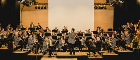 An orchestra is performing on a stage. A man is standing in the center and gesturing while playing the bassoon. Many musicians are sitting on chairs and playing various instruments. A white screen is behind them, and spotlights are mounted on the ceiling.