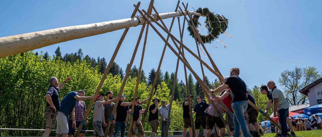 Eine Gruppe von Menschen hält einen langen Holzstab und bildet damit eine Struktur. Sie stehen auf einer offenen Wiese mit grünem Gras. Einige von ihnen tragen Hüte, Brillen und Turnschuhe. In der Ferne gibt es Bäume und einen Zaun. Der Himmel ist klar und blau.