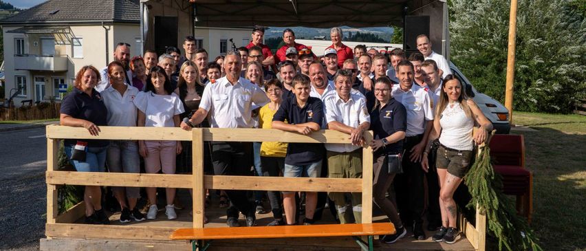 Eine große Gruppe von Menschen posiert für ein Foto vor einem Holzzaun, einige halten ein Mikrofon. Dahinter befindet sich eine Bühne mit einem Vordach.