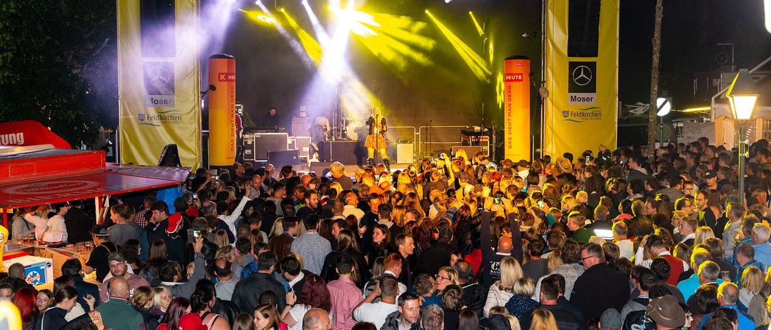 Ein Open-Air-Konzert bei Nacht mit gelbem Bühnenhintergrund und großem Publikum. Helle gelbe und lila Lichter beleuchten die Bühne, auf der eine Band auftritt.