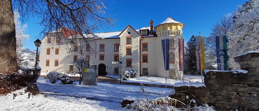 Ein großes weißes Gebäude mit rotem Rand und einem verschneiten Turm, umgeben von schneebedeckten Sträuchern und Bäumen an einem sonnigen Tag.