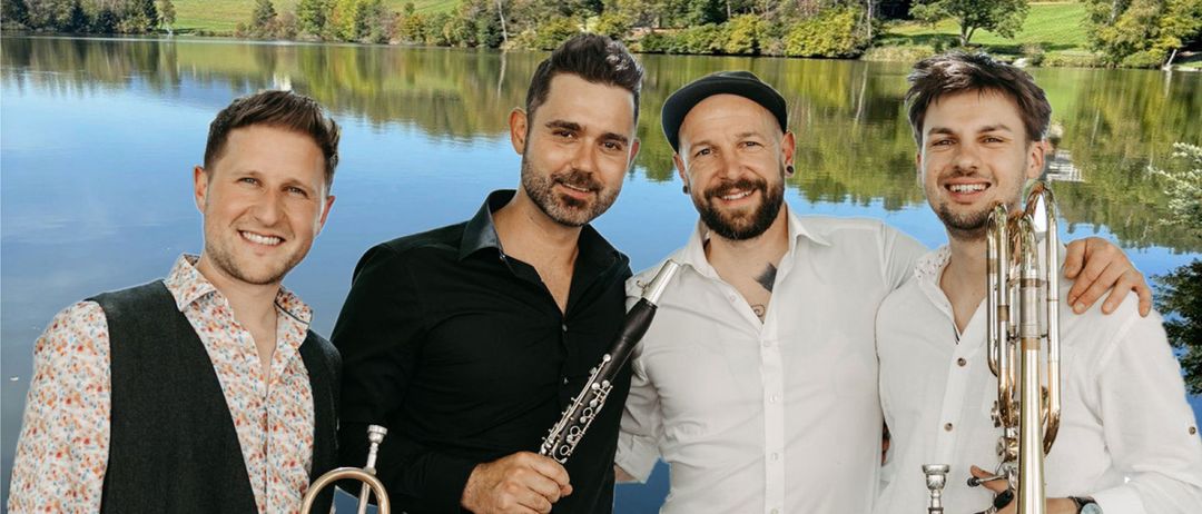 Vier Männer stehen am See mit Musikinstrumenten, lächeln und posieren für ein Foto. Dahinter spiegeln sich Bäume und Berge im ruhigen Wasser.