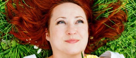 Eine Frau mit roten Haaren liegt auf grünem Gras mit gelben Blumen und lächelt. Sie trägt ein weißes Hemd. Das Sonnenlicht hebt ihre Haare und Haut hervor.