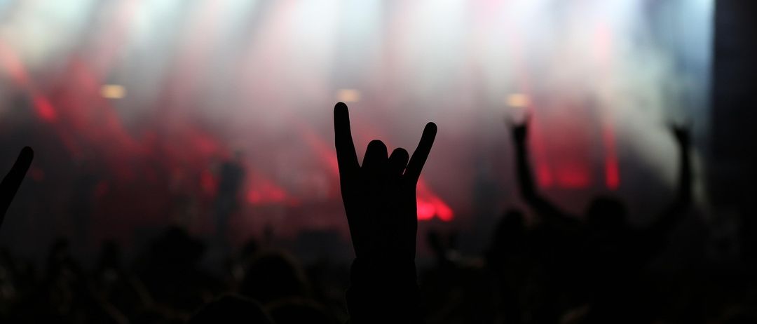 Silhouette eines Rockkonzerts mit erhobenen Händen in einer Rockgeste, rote und weiße Bühnenlichter beleuchten die Szene.
