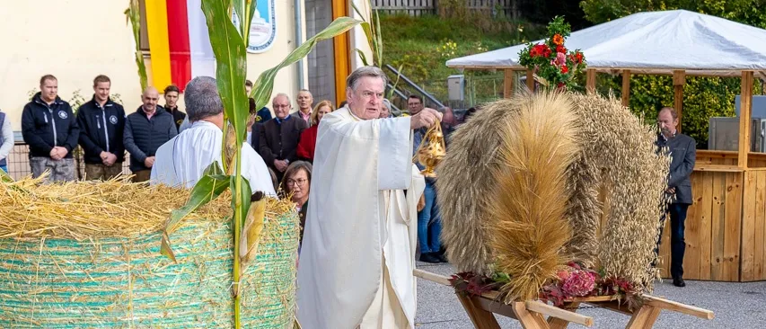Ein älterer Priester segnet ein großes Bündel Weizen bei einer Zeremonie im Freien. Menschen stehen um ihn herum. Es gibt Zelte und Blumen in der Nähe.