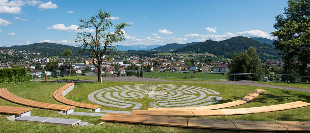 Ein runder Garten mit einem Labyrinth in der Mitte, umgeben von Holzbänken. In der Ferne befindet sich eine Stadt mit vielen Gebäuden und Bergen.