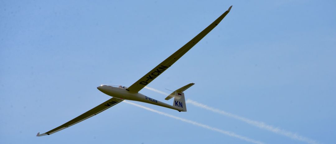A white glider with 'D-KCKN' on the wing and 'KN' on the tail flies in the sky, leaving white trails.