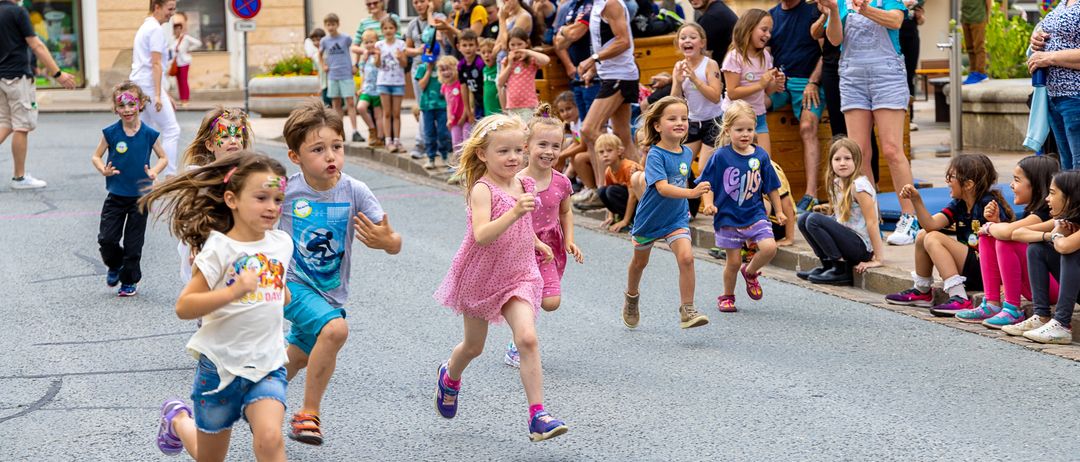 Children run in a street race with spectators cheering them on. Some wear sneakers, others sandals, all are excited. Buildings and trees line the street.