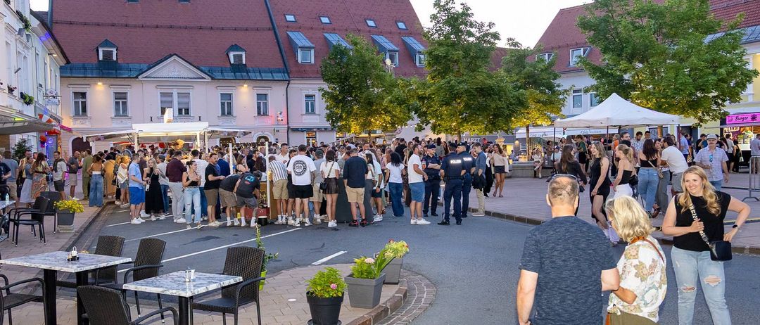 Eine überfüllte Straße mit wartenden Menschen in einer Schlange, umgeben von Bäumen und Gebäuden, mit anwesenden Polizeibeamten.