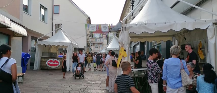 Ein geschäftiger Straßenmarkt mit Menschen, die unter weißen Zelten gehen und einkaufen. Gebäude säumen die Straße mit Vordächern und Schildern. Ein Junge hält ein buntes Kuscheltier.