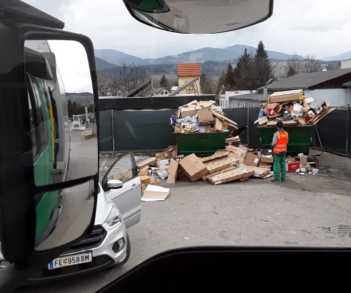 Ein Arbeiter in einer reflektierenden Weste und grünen Hose steht neben zwei großen Müllcontainern, die mit Pappkartons und anderem Müll gefüllt sind, auf einem Parkplatz.