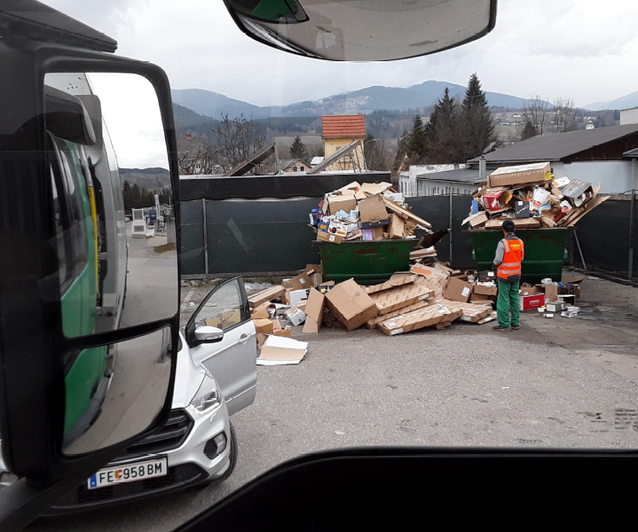 Ein Arbeiter in einer reflektierenden Weste und grünen Hose steht neben zwei großen Müllcontainern, die mit Pappkartons und anderem Müll gefüllt sind, auf einem Parkplatz.