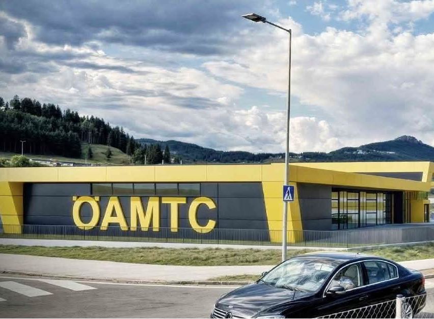 Ein schwarzes Auto fährt an einem Gebäude mit gelben und grauen Wänden vorbei, auf dem der Schriftzug 'OAMTCC' steht. Das Gebäude hat Glasfenster und ist von einem Zaun umgeben. Im Hintergrund befinden sich Bäume und Berge.