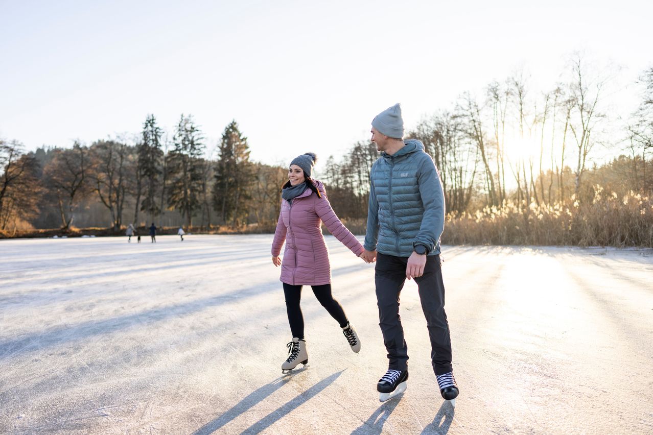 Ein Paar beim Eislaufen auf einem gefrorenen See mit Bäumen im Hintergrund und anderen Eisläufern in der Ferne.
