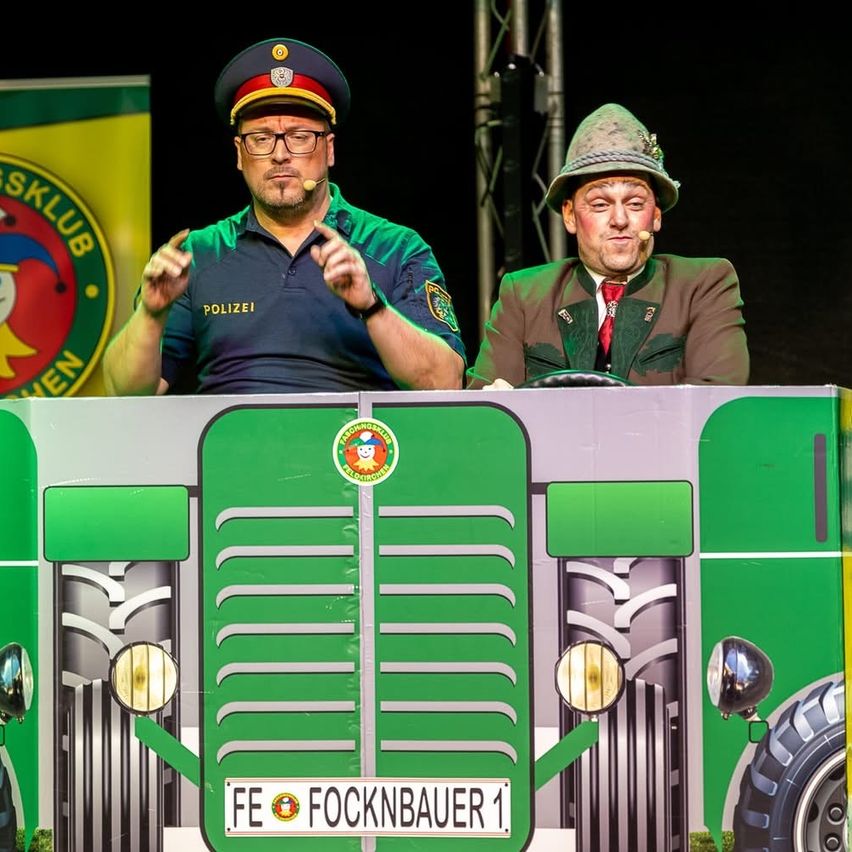 Zwei Männer auf der Bühne, einer in Polizeiuniform und der andere in einem grünen Hut, sitzen hinter einer grün lackierten Traktor-ähnlichen Struktur mit dem Nummernschild 'FE FOCKNBAUER 1'.