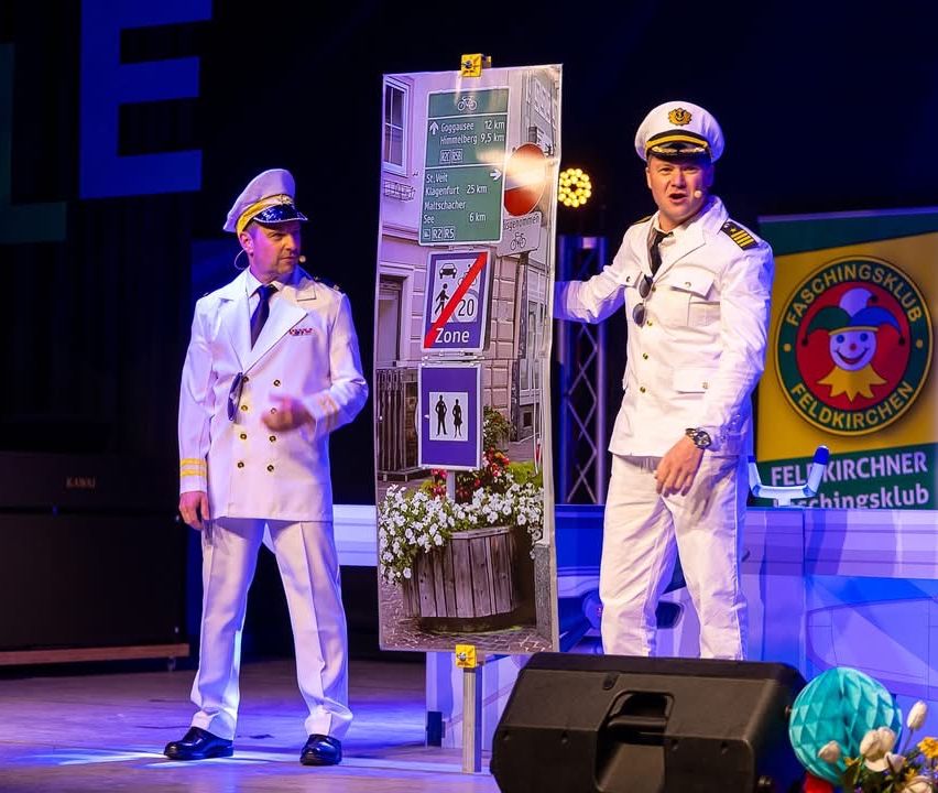 Zwei Männer in Marineuniformen stehen auf einer Bühne, einer gestikuliert. Ein großes Poster mit Straßenschildern und einem 20er-Zonenschild steht hinter ihnen. Der Hintergrund ist dunkel mit einem Scheinwerfer.