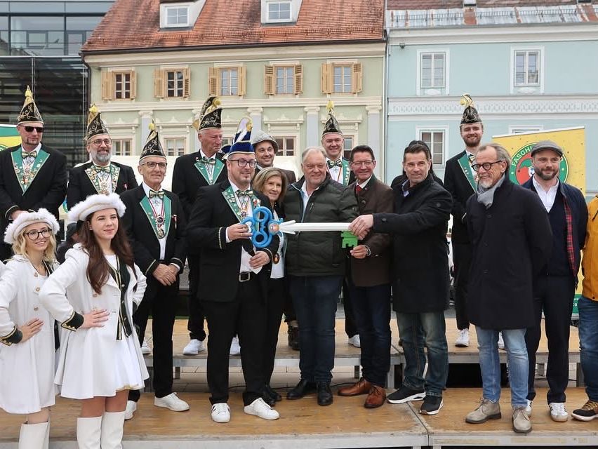 Eine Gruppe von Menschen in formeller Kleidung steht auf einer Bühne und hält einen großen Schlüssel. Einige tragen Hüte, Brillen und Halsketten. Eine Frau in Weiß steht vorne. Im Hintergrund befinden sich Gebäude.