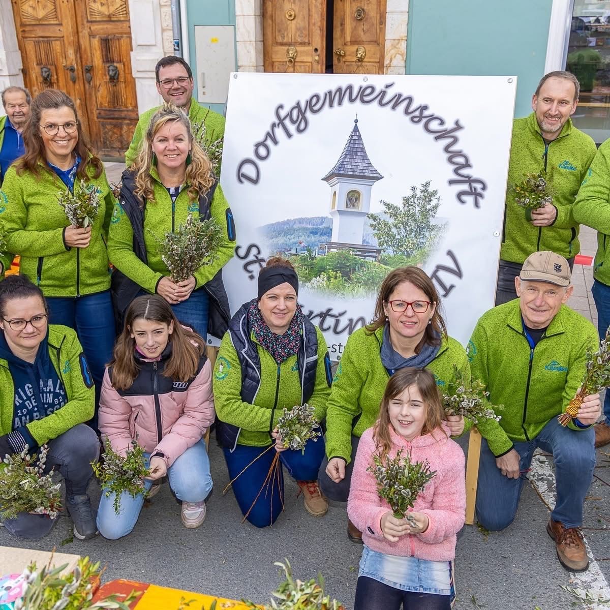 A group of people in green jackets pose with flowers in front of a sign that reads Dorfgemeinschaftshaus.