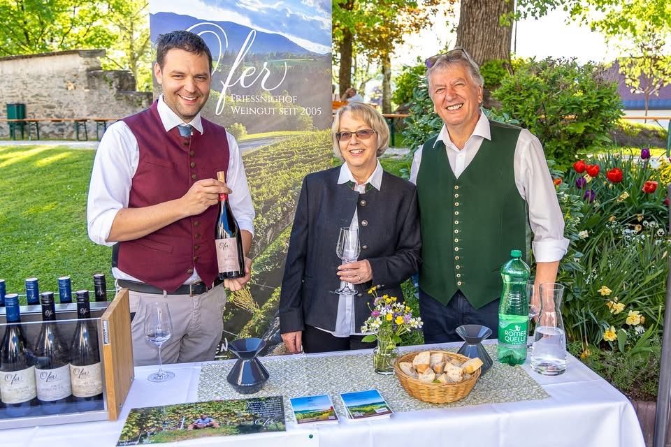 Drei Personen stehen bei einem Tisch mit Weinflaschen, Gläsern und einem Korb mit Brot und lächeln für ein Foto.