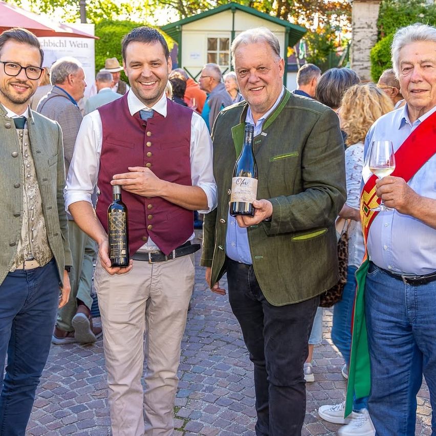 Vier Männer stehen lächelnd zusammen auf einer Kopfsteinpflasterstraße. Einer hält eine Weinflasche, ein anderer ein Weinglas. Sie tragen traditionelle deutsche Kleidung.