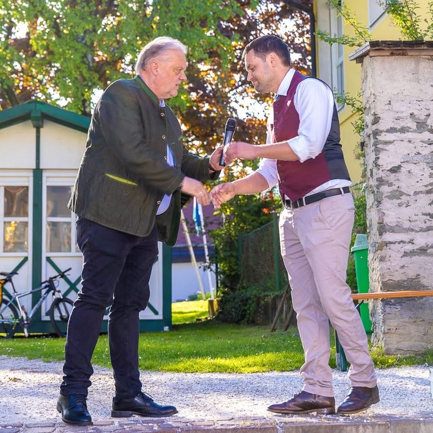 Zwei Männer stehen auf einem Gehweg, einer hält ein Mikrofon und der andere schüttelt Hände. Der ältere Mann trägt eine grüne Jacke. Hinter ihnen ist ein Haus mit grünem Vordach und Fahrrädern zu sehen.