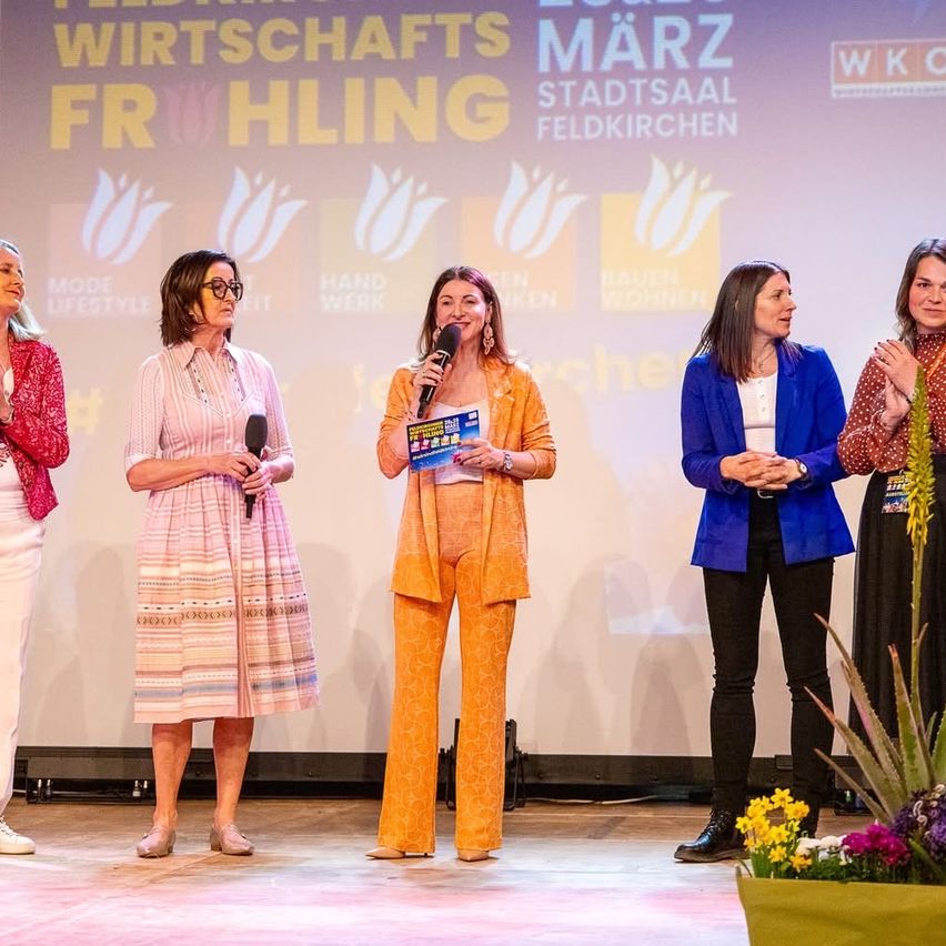 Fünf Frauen stehen mit Mikrofonen auf der Bühne, eine hält ein Buch. Hinter ihnen befindet sich ein Bildschirm mit Text über Wirtschafts und Stadtsaal Feldkirchen.