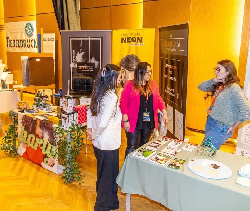Drei Frauen stehen vor einem Tisch mit Büchern bei einer Indoor-Veranstaltung, in der Nähe eines Plakats mit einem Mannequin.