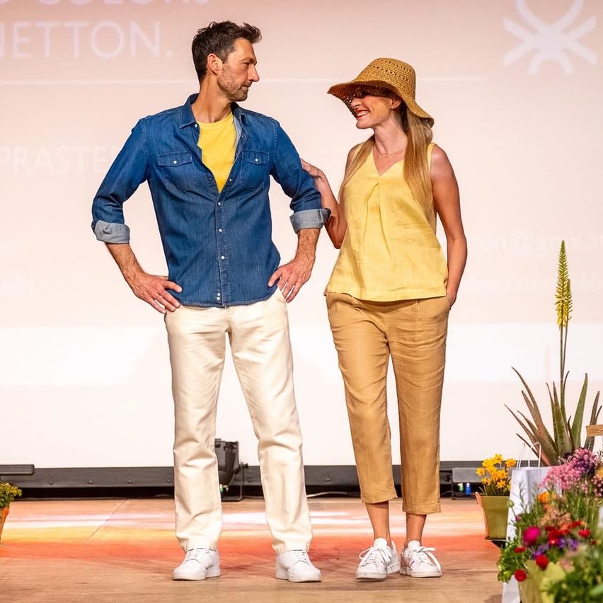 Ein Mann und eine Frau stehen lächelnd auf der Bühne und schauen sich an. Der Mann trägt ein Jeanshemd und beige Hosen. Die Frau trägt ein gelbes Oberteil und Hosen.