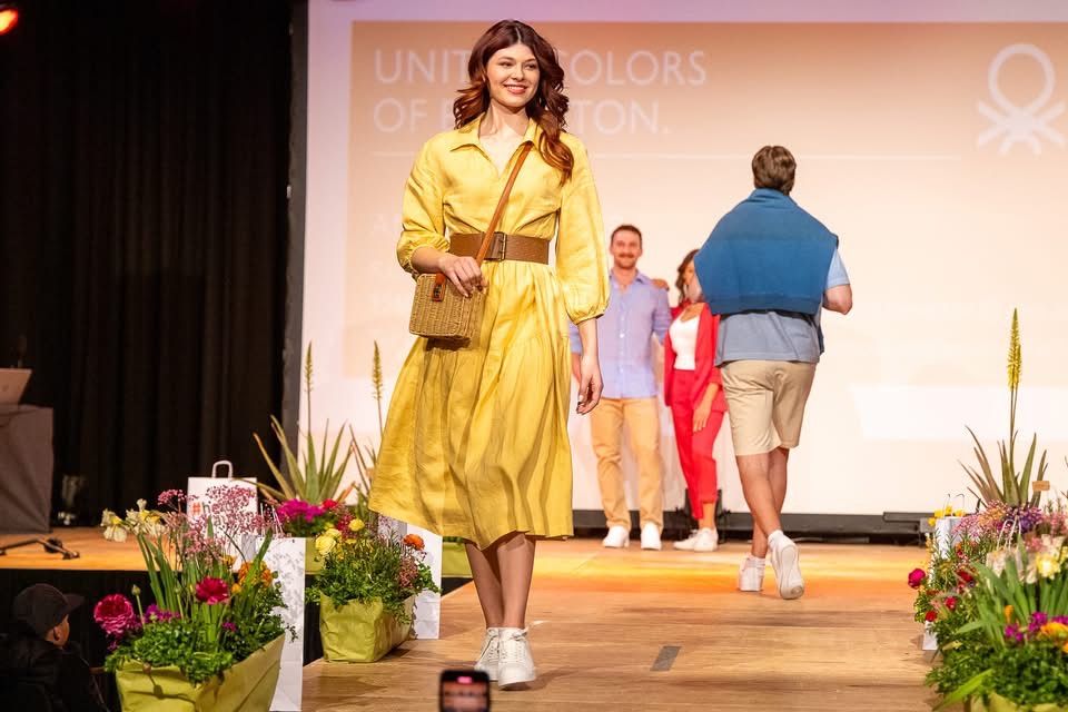 Eine Frau läuft auf einer Bühne mit Blumen im Vordergrund. Dahinter laufen ein Mann und eine Frau gemeinsam. Eine Projektionswand zeigt 'UNITY OF BOSTON.'