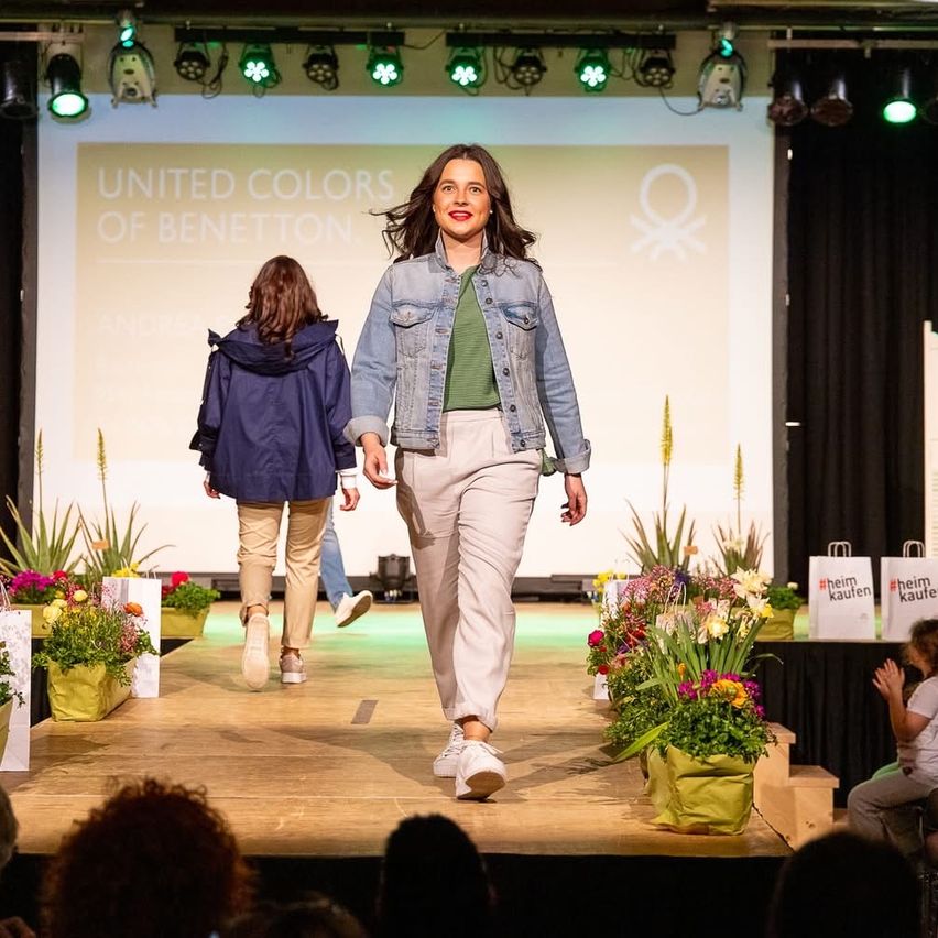 Eine Modenschau findet statt. Eine Frau mit Blumen geht auf die Bühne. Ein Schild zeigt United Colors of Benetton. Ein junges Mädchen folgt ihr.