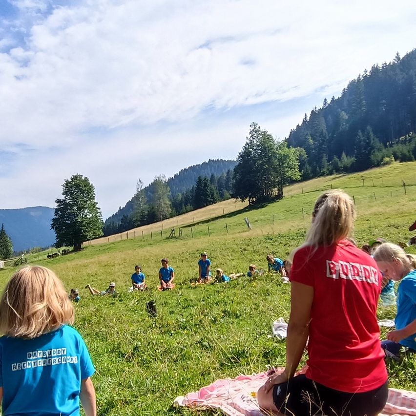 A group of children and adults are sitting on a grassy field, possibly in a camp, with a woman sitting on a blanket in front of them. The area is surrounded by trees and mountains.